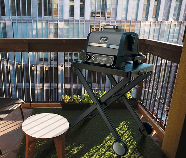 Barbecue su supporto pieghevole con ruote, sgabello rotondo, ringhiera e luci su un balcone. Edificio in background.