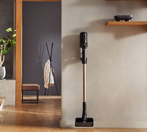 Aspirapolvere verticale nera e oro accanto al muro beige. In secondo piano, appendiabiti nero e panca marrone in stanza grigia.