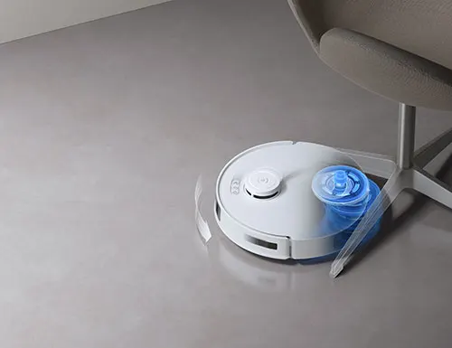 Aspirapolvere robot bianco vicino a una sedia su un pavimento grigio. La spazzola laterale � di colore blu.