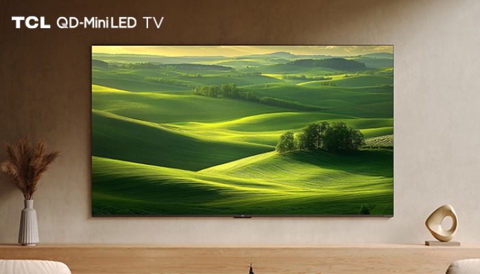 Un televisore TCL QD-Mini LED TV montato a parete sopra un mobile in legno chiaro. Sullo schermo è visualizzato un paesaggio di colline verdi illuminate dal sole.