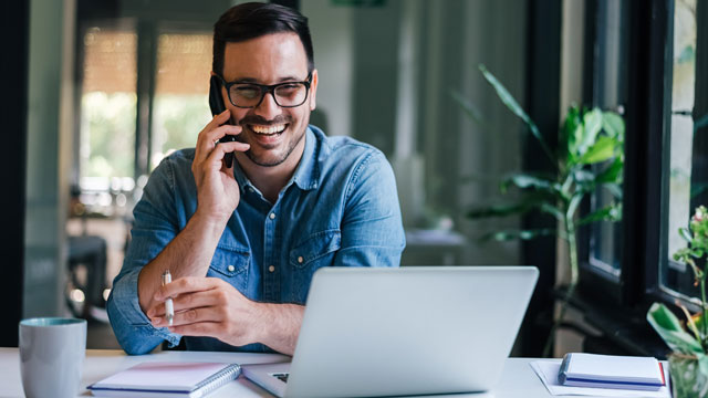 Uomo sorridente al telefono con portatile, taccuini e tazza su scrivania bianca. Finestra con piante sullo sfondo.