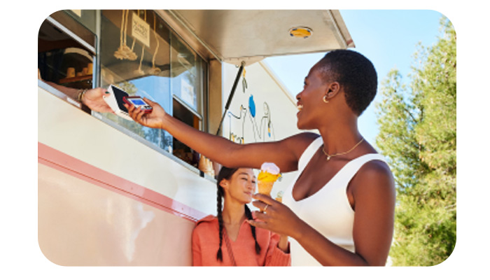 Donna paga con cellulare al food truck. Un'altra donna con trecce tiene un gelato. Alberi sullo sfondo.