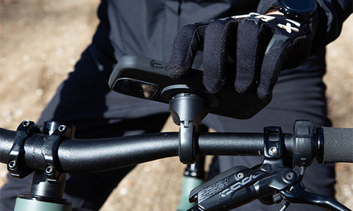 Primo piano del manubrio di una bicicletta con una mano guantata sopra. Si vedono i freni e un supporto per telefono.