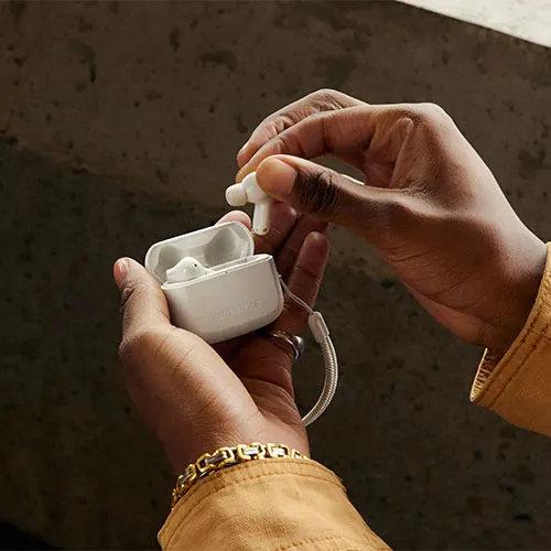 Mani tengono auricolari bianchi e custodia aperta. Si vedono un braccialetto dorato e parte di una giacca marrone.