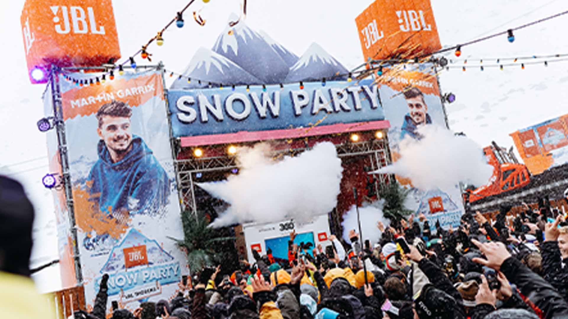 Folla a un evento all'aperto "Snow Party" con palco, montagne gonfiabili e striscioni JBL.