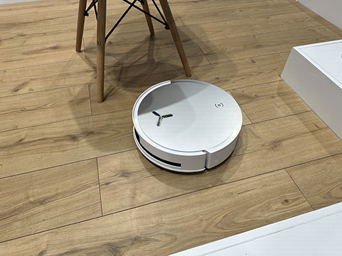 Aspirapolvere robot bianco sul pavimento in legno vicino a una sedia di legno e una scatola bianca.