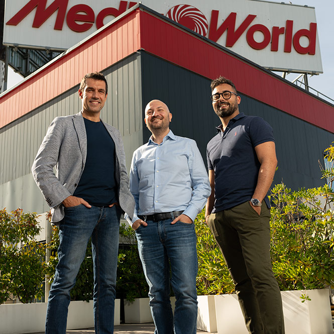 Tre uomini posano davanti a un edificio con un'insegna "Media World". Indossano abiti casual e sorridono. Arbusti sullo sfondo.