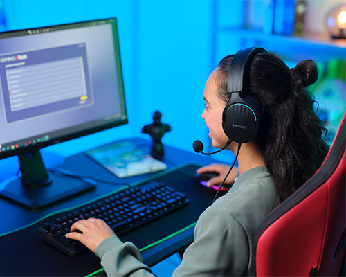 Ragazza con cuffie gioca al computer su sedia rossa. Sul tavolo tastiera, mouse e monitor. Sfondo blu.