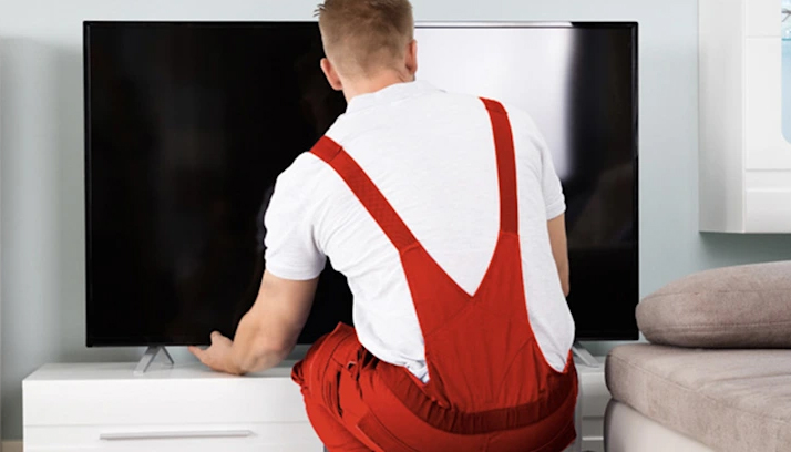 Uomo di spalle in salopette rossa che sistema una TV nera su un mobile bianco in una stanza con divano beige.