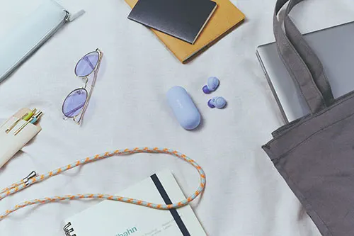 Borsa grigia, laptop, occhiali, auricolari blu, custodia, portafoglio con penne, quaderni e cordino su sfondo chiaro.