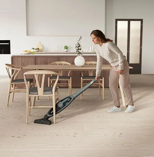 Donna aspira il pavimento di legno con aspirapolvere grigio. Tavolo e sedie in legno sullo sfondo.