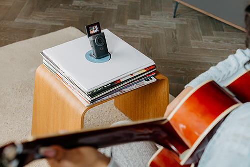 Una fotocamera nera su una pila di dischi in vinile bianchi su un tavolino di legno. Un uomo suona la chitarra in primo piano.
