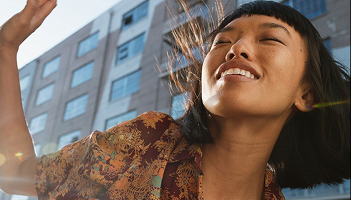 Donna sorride, occhi chiusi, indossa una camicia a motivi floreali. Dietro, un edificio moderno con finestre. Mano alzata in alto.