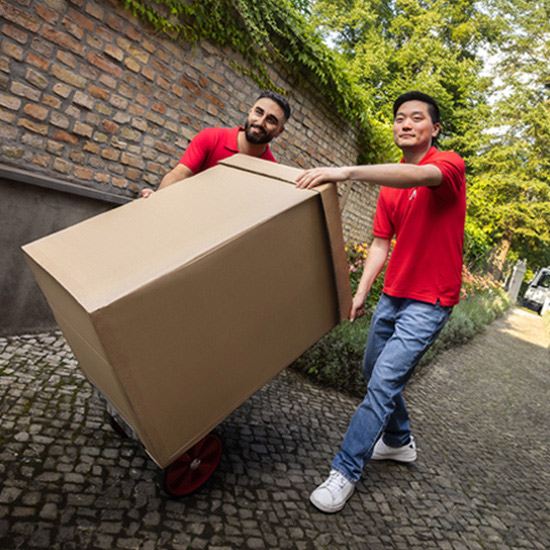 Due persone in magliette rosse trasportano una grande scatola di cartone su un carrello su una strada di ciottoli vicino a un muro di mattoni.