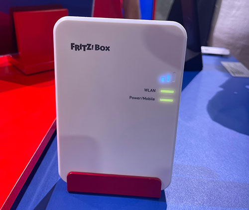 Un router bianco Fritz!Box su supporto rosso, con spie WLAN blu e Power/Mobile verdi. Sfondo rosso e blu.