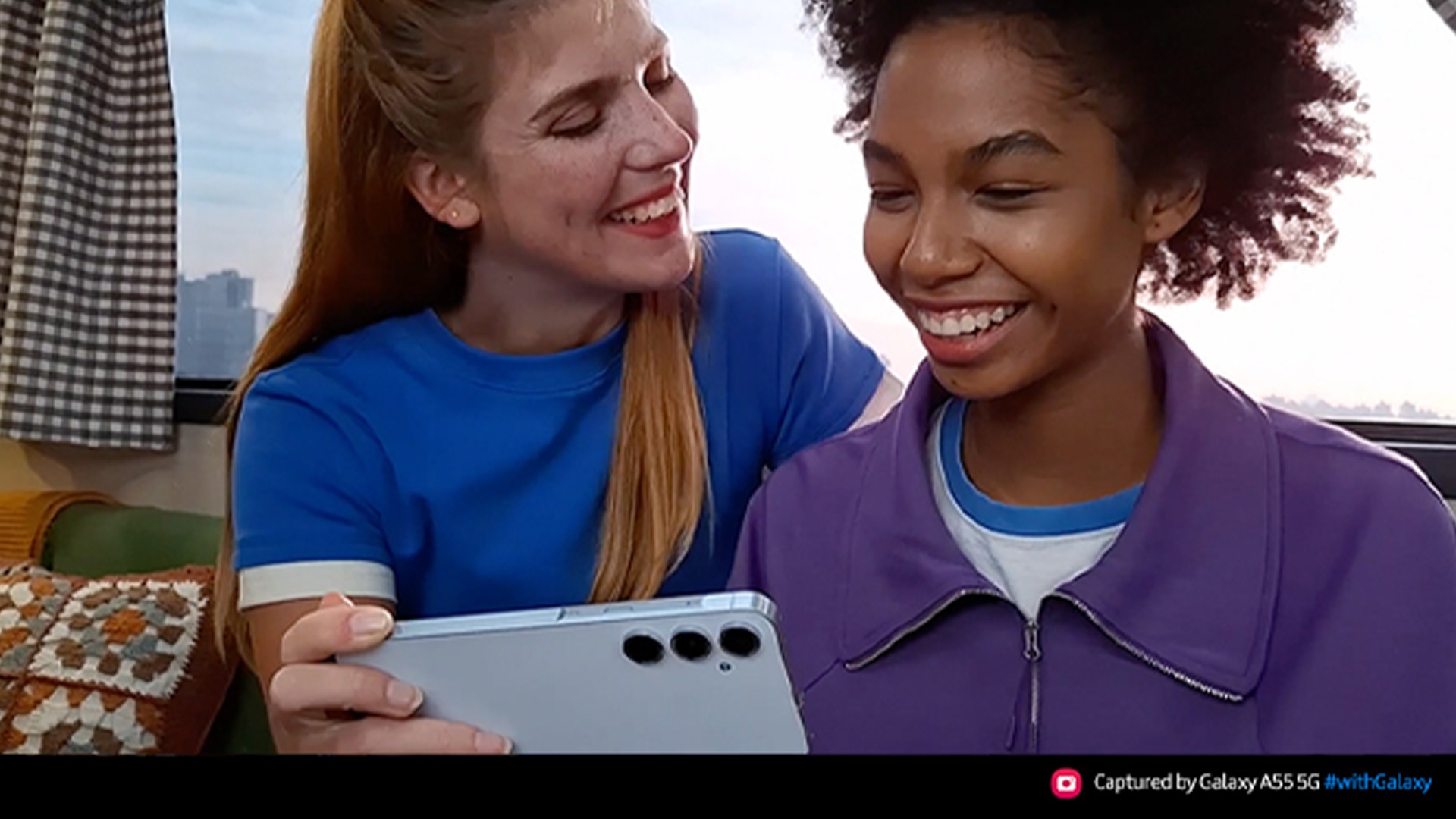 Due donne sorridono mentre guardano un telefono argento. La donna a sinistra indossa una maglietta blu. L'altra indossa una giacca viola.