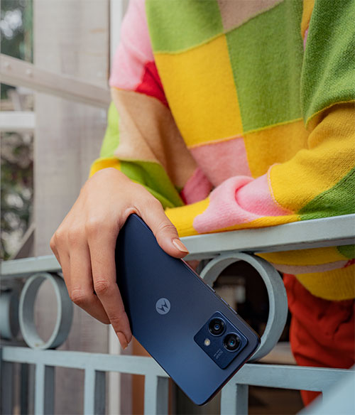 Mano che tiene un telefono blu con doppia fotocamera, su una ringhiera grigia. Indossa una maglia a scacchi colorata.