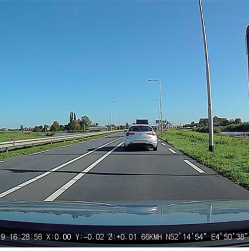 Strada asfaltata con auto bianca in lontananza sotto cielo sereno. Aiuole verdi ai lati della strada.