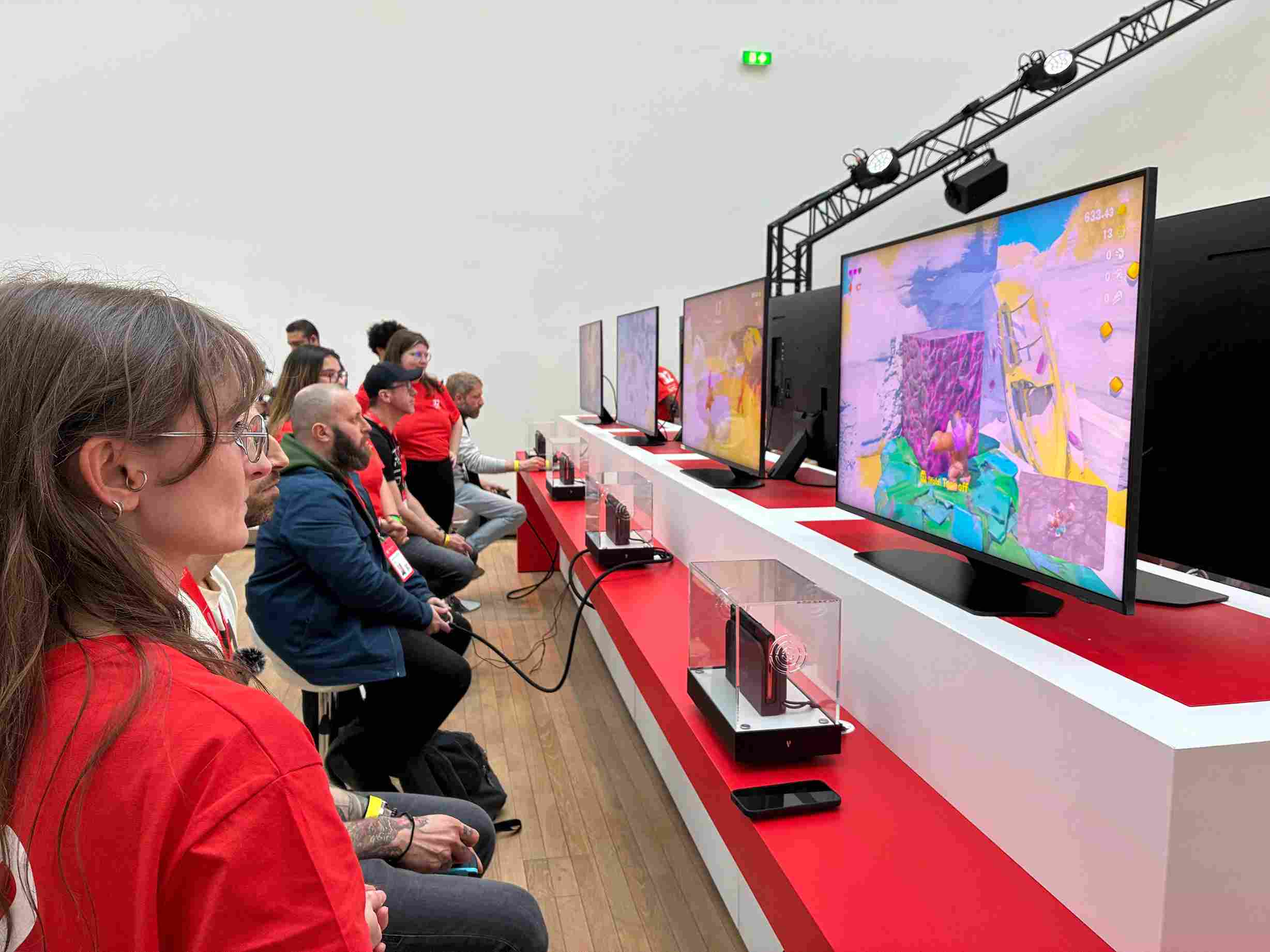 Persone giocano a videogiochi su schermi multipli. Console in vetrine su un lungo tavolo bianco e rosso.