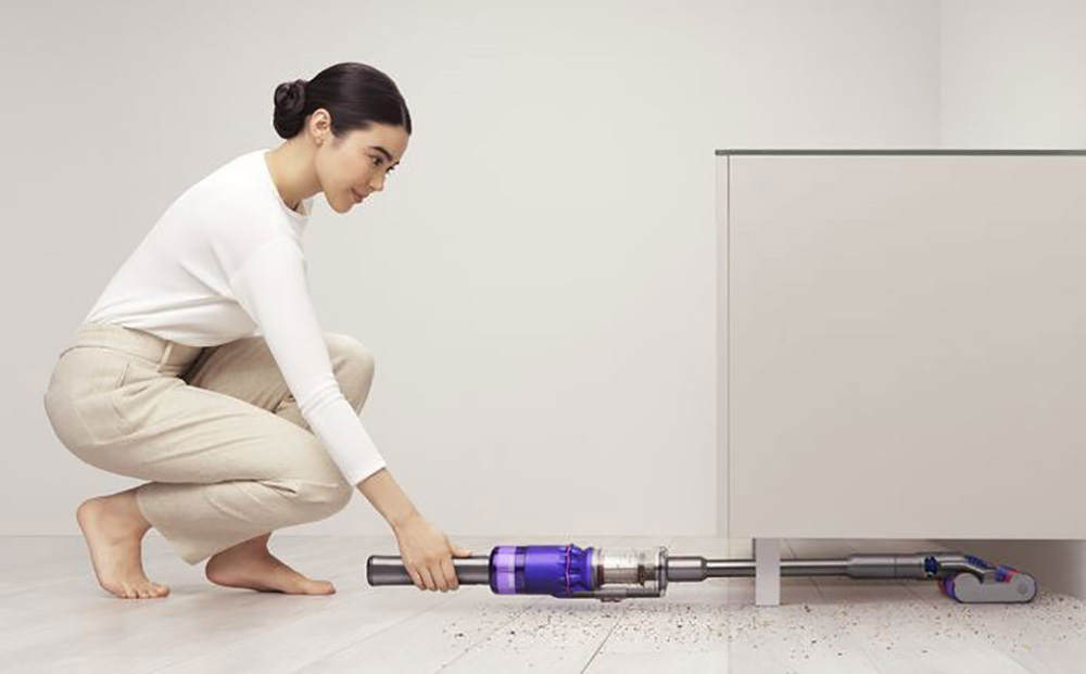 Donna accovacciata usa aspirapolvere viola e argento sotto un mobile bianco. Il pavimento � chiaro.