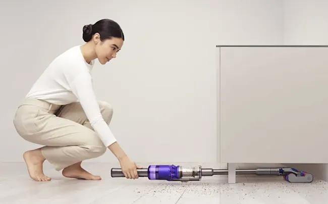 Donna accovacciata usa aspirapolvere viola e argento sotto un mobile bianco. Il pavimento � chiaro.
