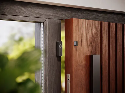 Porta scorrevole in legno marrone con maniglia nera, accanto a una finestra con cornice scura e vegetazione sfocata.