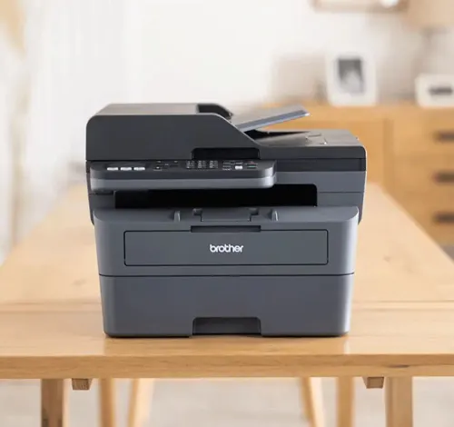 Stampante Brother grigia su tavolo di legno. Sullo sfondo, un mobile e delle foto.