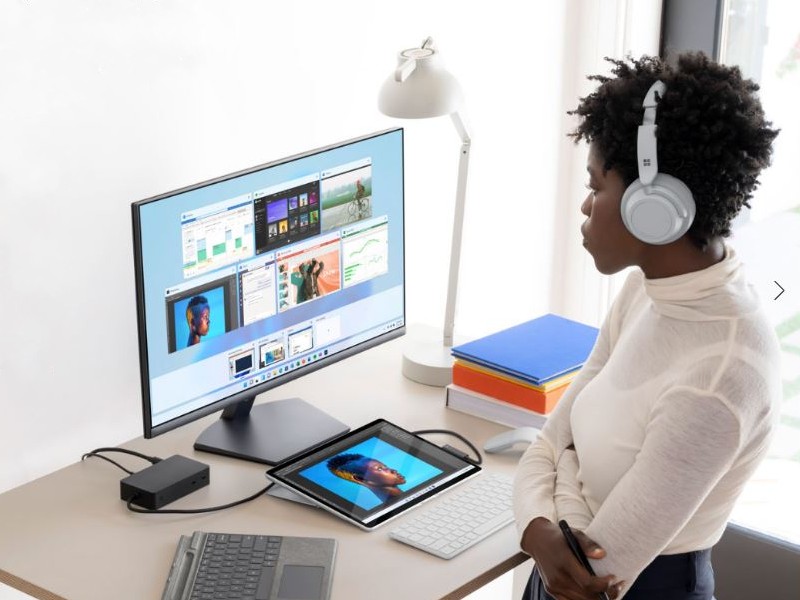 Donna con cuffie guarda monitor con finestre aperte, tablet, tastiera su scrivania. Libri e lampada sullo sfondo.