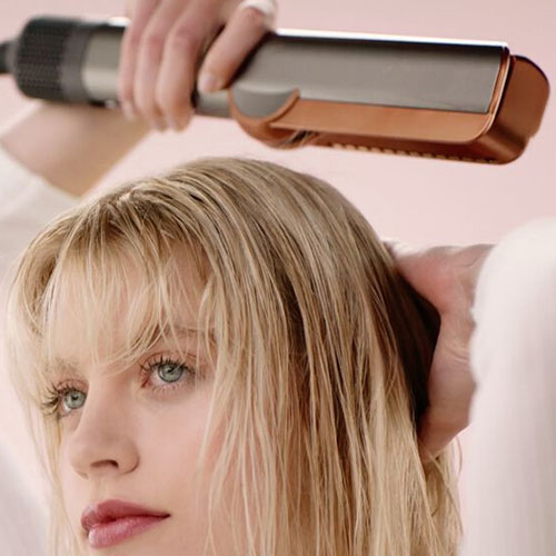 Donna che riceve una piega con una piastra per capelli color bronzo. Le mani tengono la piastra sopra la sua testa.