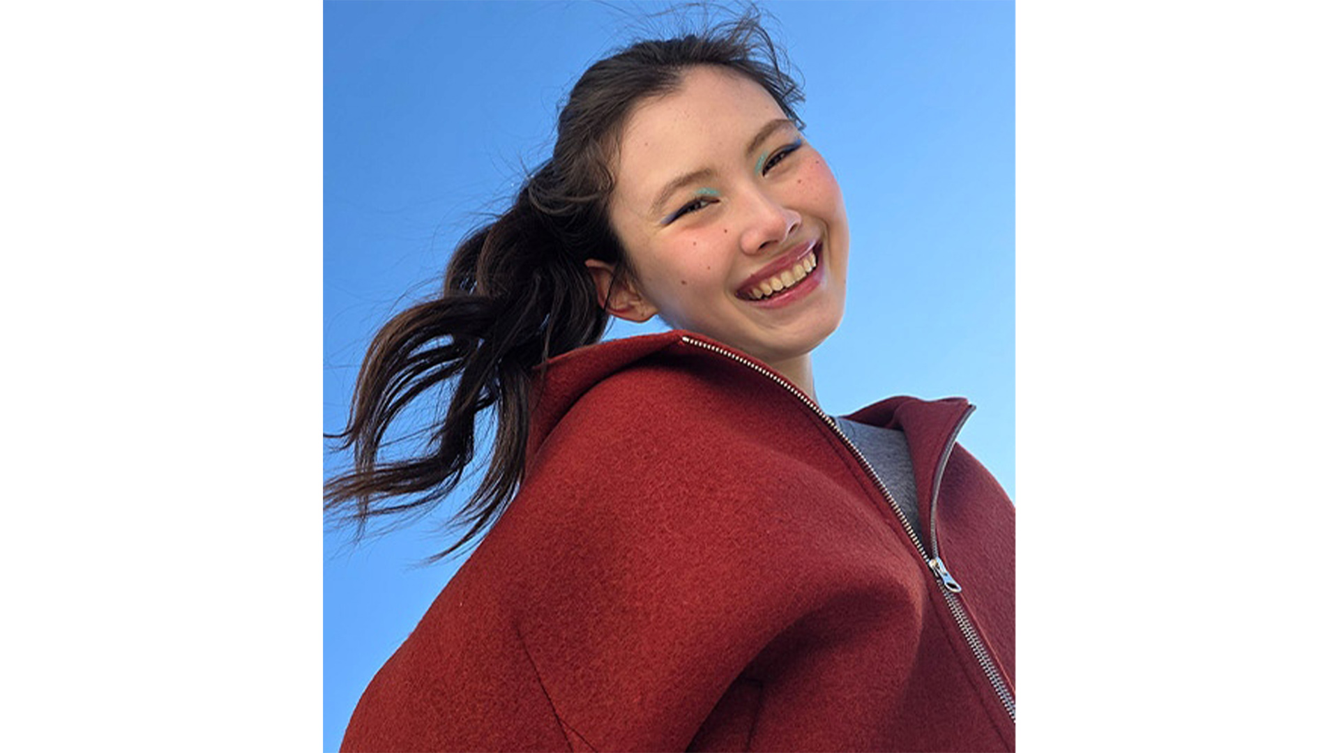 Ragazza sorridente con giacca rossa e maglietta grigia. Capelli scuri raccolti, cielo azzurro sullo sfondo.