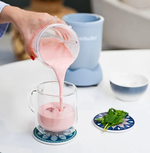 Su un tavolo bianco, una mano versa un frullato rosa in un bicchiere. Un frullatore blu, una ciotola bianca e verdure sono visibili.