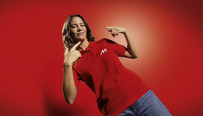 Donna su sfondo rosso che indica la maglietta rossa con il logo bianco e indossa jeans blu.