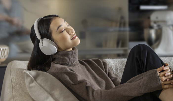 Donna con cuffie bianche, maglione marrone e pantaloni neri, seduta su un divano chiaro. Sullo sfondo si intravede una cucina.
