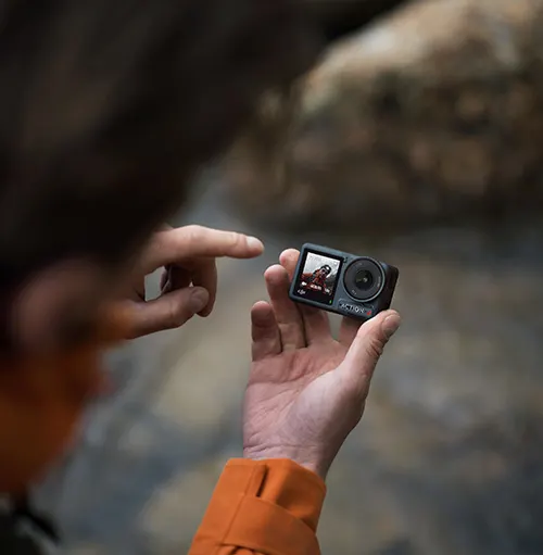 Uomo con giacca arancione che tiene una action camera nera, puntando il dito verso lo schermo. Sfondo sfocato con rocce.
