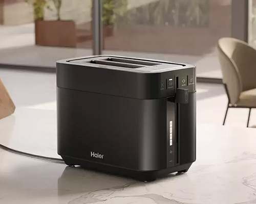 Tostapane nero su un piano chiaro, marca Haier visibile. Sullo sfondo, una sedia e una finestra con piante.