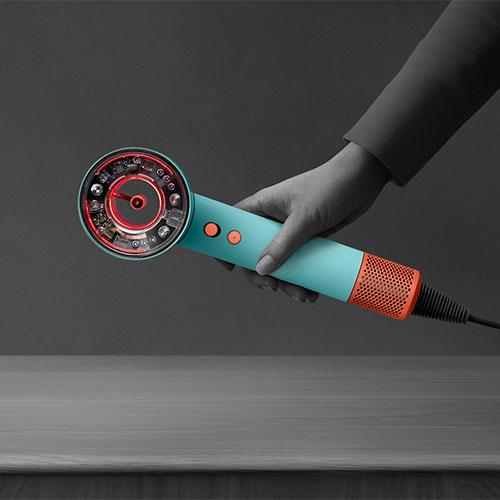 Una mano tiene un asciugacapelli azzurro e arancione su una superficie grigia, con uno sfondo grigio scuro.