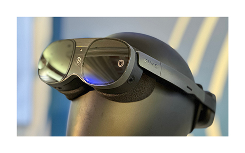 Occhiali VR neri su supporto. Le lenti riflettono la luce. Fascia regolabile. Sfondo sfumato blu e bianco.