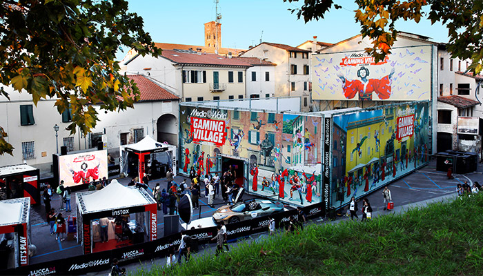 Evento all'aperto con stands e folla di persone. Edifici sullo sfondo e cartelloni pubblicitari visibili.