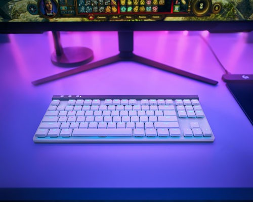 Tavolo illuminato di viola con tastiera bianca, monitor e mouse neri. Lo schermo mostra un gioco.