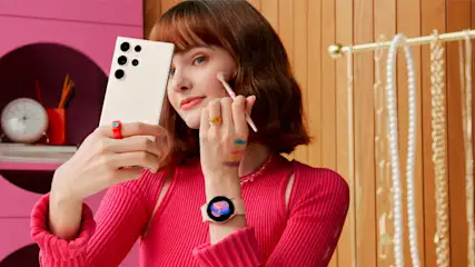 Donna con pennello cosmetico e smartphone bianco con tre obiettivi. Indossa un orologio e gioielli. Dietro, scaffale rosa e appendiabiti oro.