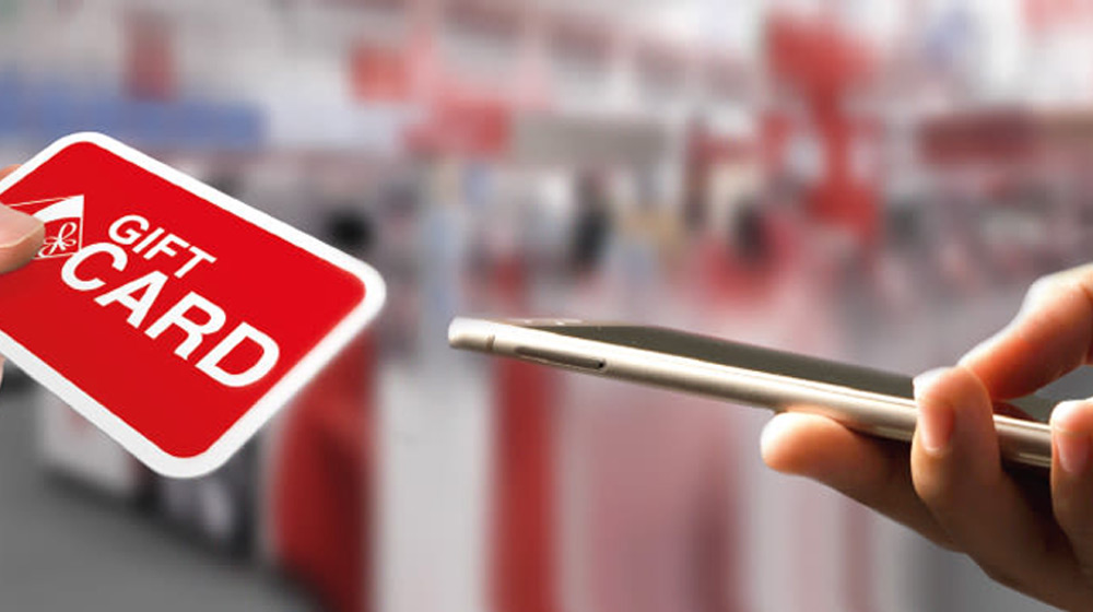 Una mano tiene una carta regalo rossa con scritte bianche, mentre un'altra mano tiene un telefono.