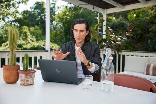 Uomo con portatile a un tavolo con cactus e bottiglia d'acqua. Dietro, vegetazione e balcone con ringhiera bianca.