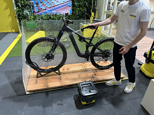 Bicicletta nera su piattaforma di legno dietro una barriera trasparente. Una persona la pulisce con un'idropulitrice gialla.