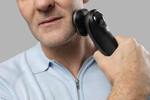 Uomo che si rade con rasoio elettrico nero su sfondo grigio. Indossa una camicia azzurra con cerniera argentata.