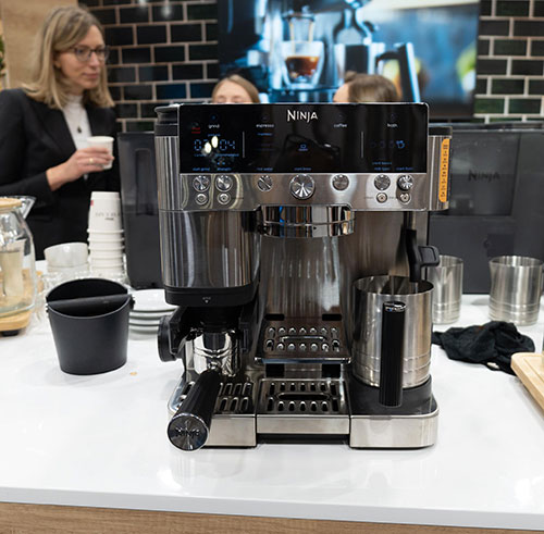 Macchina da caffè Ninja in acciaio su bancone bianco, con tazza, tazzine e accessori. Sullo sfondo, parete di mattoni.