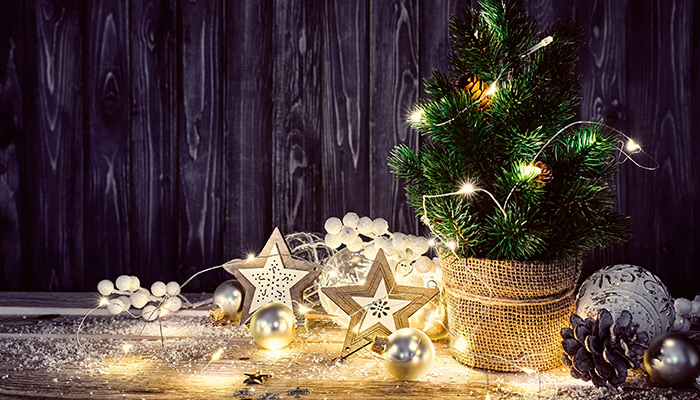 Decorazioni natalizie su legno: albero illuminato, stelle decorative, palline argentate e pigna. Luci a batteria sparse.