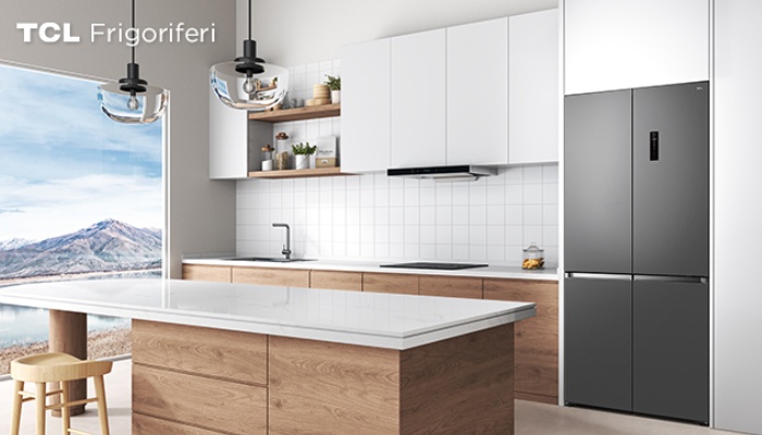 Un frigorifero quattro porte TCL color grigio scuro incassato nei mobili di una cucina moderna e luminosa, caratterizzata da un'isola centrale in legno con piano in marmo bianco.