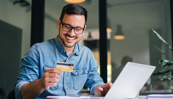 Uomo sorridente con camicia di jeans e occhiali che tiene una carta di credito davanti a un laptop. Pianta sullo sfondo.