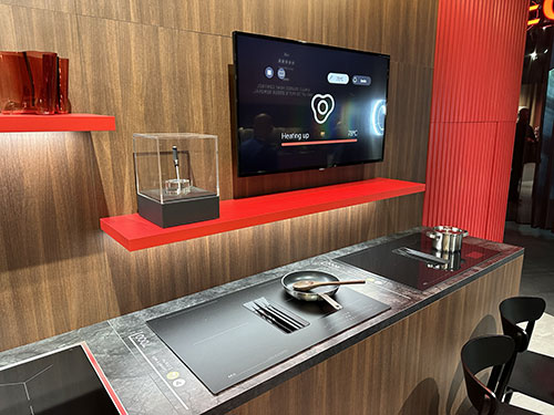 Cucina moderna con piano cottura nero, padella e utensili. Mensola rossa con TV e oggetti decorativi su sfondo in legno e colonna rossa.