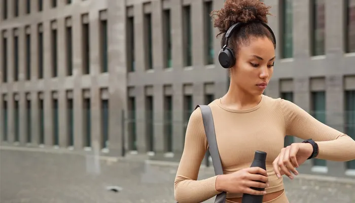 Una donna con cuffie grandi e abbigliamento sportivo beige controlla lo smartwatch al polso mentre tiene una borraccia nera, con un edificio moderno sullo sfondo.